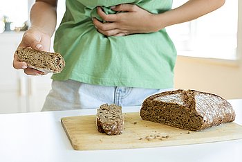 Hier ist ein Mensch zu sehen, der offensichtlich Bauchschmerzen hat. Er hält ein Stück Vollkornbrot in der Hand. Auf dem Tisch vor ihm ist der Rest des Vollkornbrots zu sehen. Bei Menschen mit Zöliakie kann Gluten nicht die einzige Ursache für Verdauungsbeschwerden sein. Es sollte auch an die Bauchspeicheldrüse gedacht werden, da eine begleitende exokrine Pankreasinsuffizienz bei Menschen mit Zöliakie möglich sein kann. 