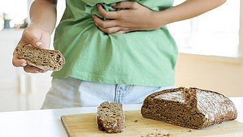 Hier ist ein Mensch zu sehen, der offensichtlich Bauchschmerzen hat. Er hält ein Stück Vollkornbrot in der Hand. Auf dem Tisch vor ihm ist der Rest des Vollkornbrots zu sehen. Bei Menschen mit Zöliakie kann Gluten nicht die einzige Ursache für Verdauungsbeschwerden sein. Es sollte auch an die Bauchspeicheldrüse gedacht werden, da eine begleitende exokrine Pankreasinsuffizienz bei Menschen mit Zöliakie möglich sein kann. 