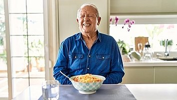 Ein älterer sitzt am Tisch und ißt Spaghetti und hält sich dabei mit schmerzverzehrtem Gesicht den Bauch.